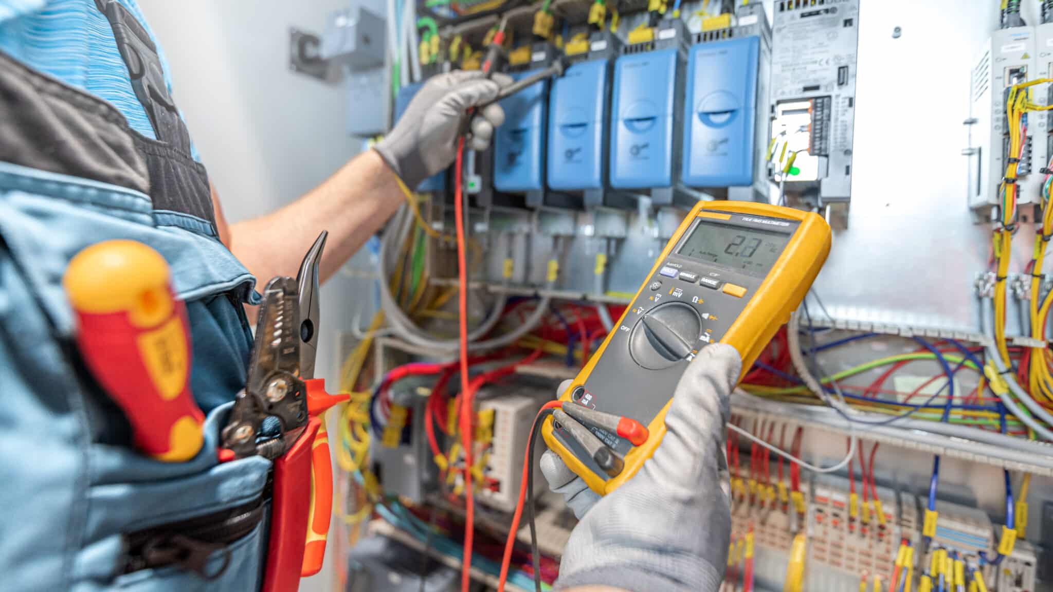 The electrician’s hand is holding a multimeter while checking the electrical wiring in the fuse box for emergency electrical repairs.